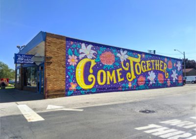 Photo of exterior public art mural with the phrase Come Together surrounded by flowers.