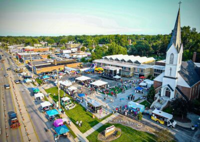 Exterior aerial view of a St. John Lutheran Church and Mayfield Road during the summer festival, Rock the Block. Tents line the street, a large stage for music is situated in the center of the parking lot, and crowds of people enjoy the festivities.